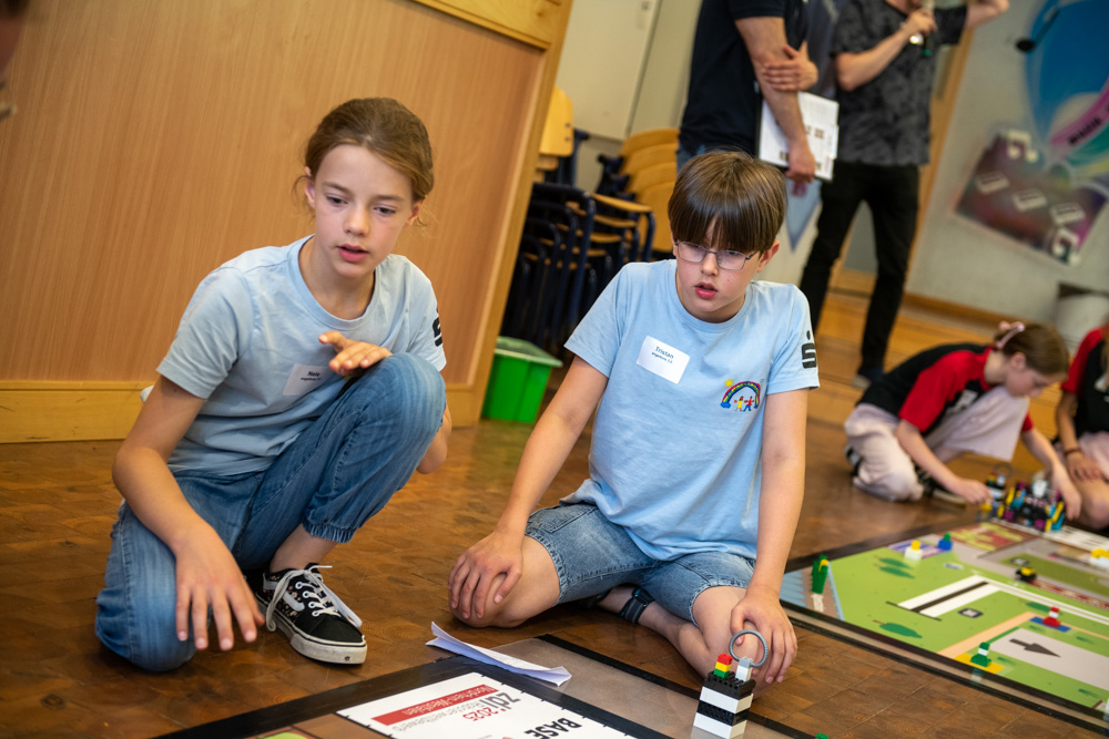 Zwei Kinder sitzen in einem Klassenzimmer auf dem Boden. Sie beschäftigen sich mit einer MINT-Aktivität und haben vor sich Bauklötze und ein Spielbrett.