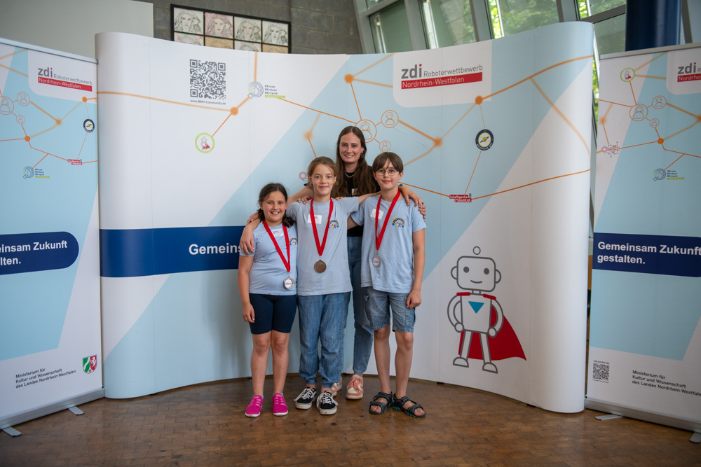 Vier Kinder mit Medaillen stehen drinnen vor einer zdi-Ausstellungskulisse, hinter ihnen steht eine Frau.