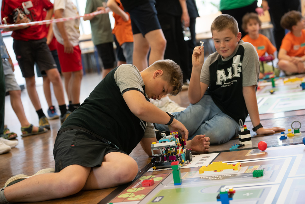 Zwei Jungen sitzen auf dem Boden und bauen und programmieren bei einem Roboterwettbewerb einen Roboter. Andere Kinder stehen im Hintergrund und schauen zu.