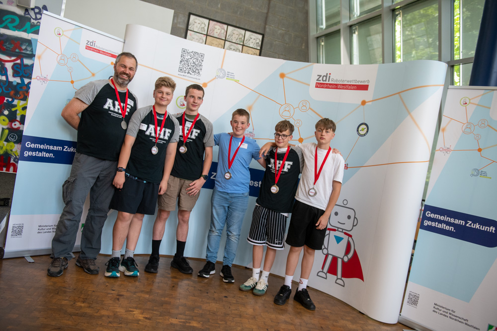 Fünf Jungen und ein Mann stehen vor einer naturwissenschaftlichen Kulisse. Sie tragen Medaillen und posieren für ein Gruppenfoto bei einer Indoor-Veranstaltung.