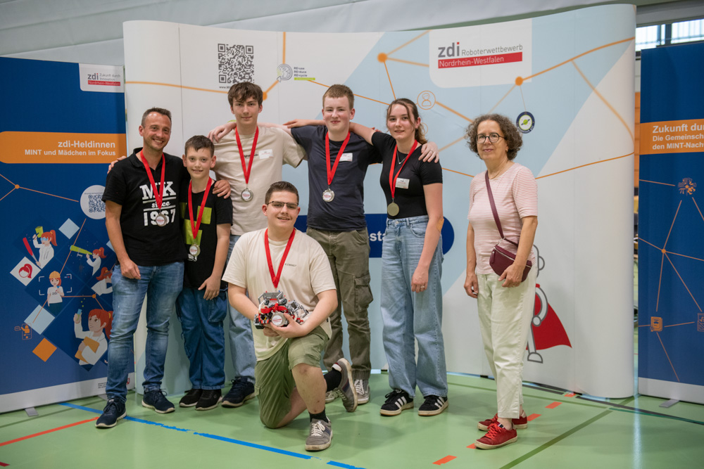 Sechs Jugendliche und ein Erwachsener stehen in einem Raum und lächeln vor Veranstaltungsbannern. Einige der jungen Leute tragen Medaillen, was darauf hindeutet, dass sie an einer wissenschaftlichen oder technischen Veranstaltung teilgenommen haben.