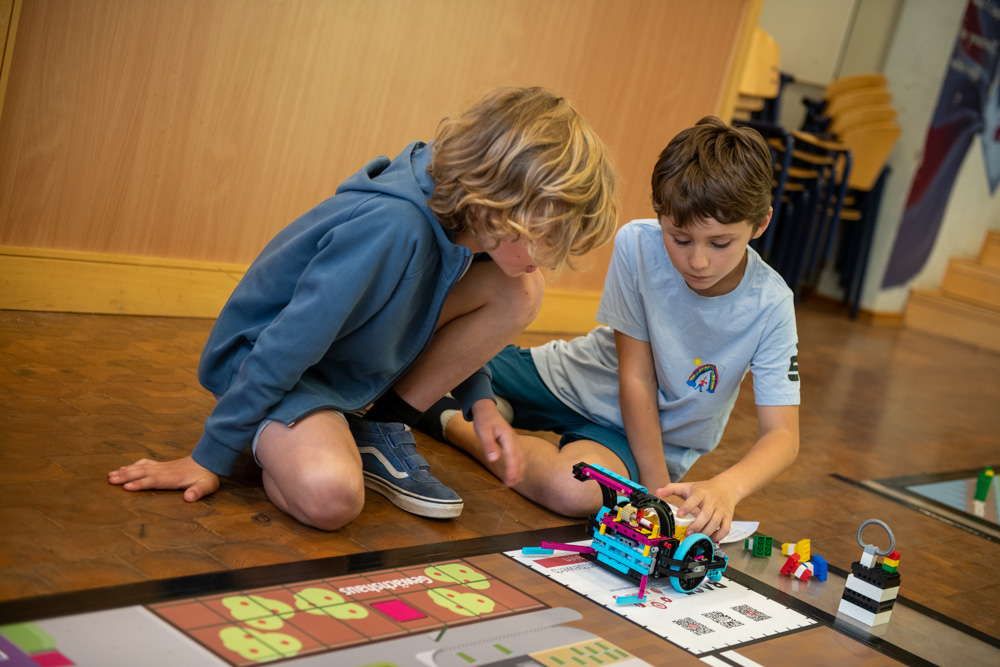 Zwei Jungen sitzen in einem Klassenzimmer auf dem Boden. Gemeinsam bauen sie auf einer Spielmatte einen bunten LEGO-Roboter zusammen.