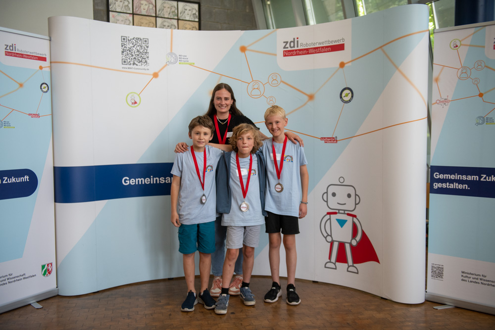 Vier Kinder mit Medaillen stehen bei einer Veranstaltung des ZDI vor einem mit Grafiken aus Wissenschaft und Technik verzierten Banner.