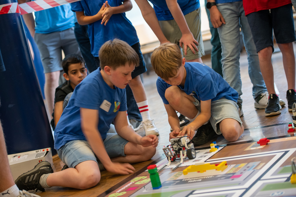 Zwei Jungen in blauen Shirts und Shorts knien auf dem Boden und arbeiten bei einem Roboterwettbewerb an einem kleinen Roboter. Andere Kinder schauen zu.