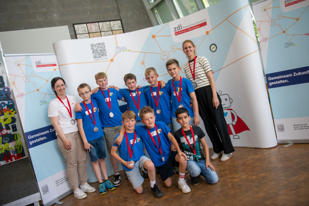 Eine Gruppe von Kindern in blauen Hemden und Medaillen steht mit zwei Erwachsenen vor einer Indoor-Veranstaltungskulisse mit naturwissenschaftlichem Thema.