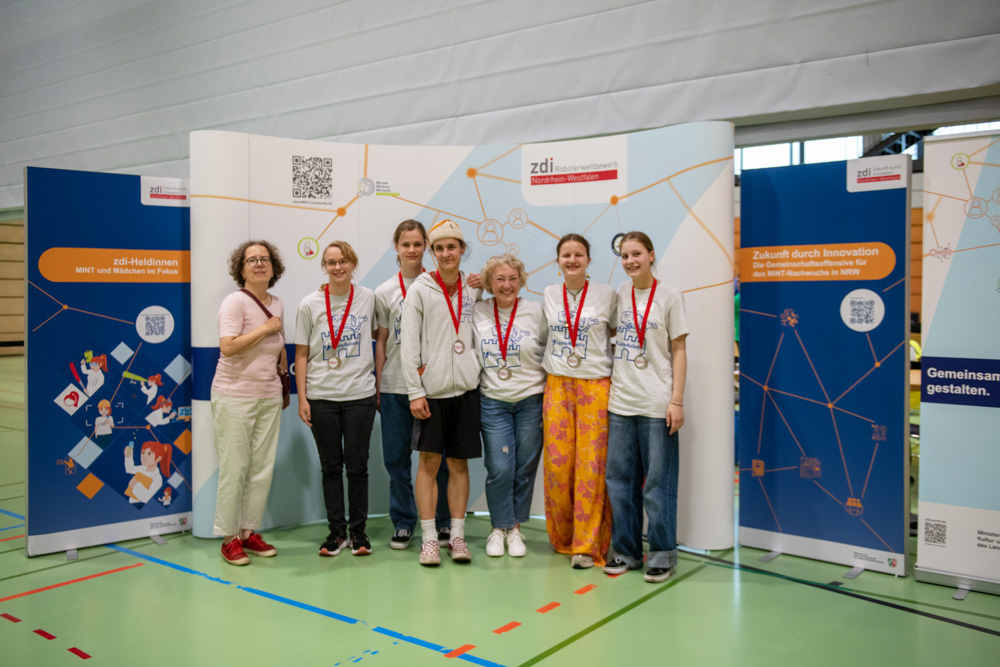 Sechs Personen in Freizeitkleidung mit Medaillen stehen drinnen vor Informationstafeln und posieren für ein Gruppenfoto.