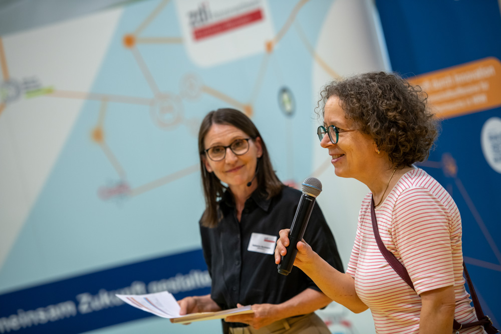Zwei Frauen stehen in einem Haus. Eine Frau hält Papiere in der Hand, die andere spricht in ein Mikrofon. Beide Frauen tragen eine Brille. Im Hintergrund ist ein unscharfes Informationsplakat zu sehen.