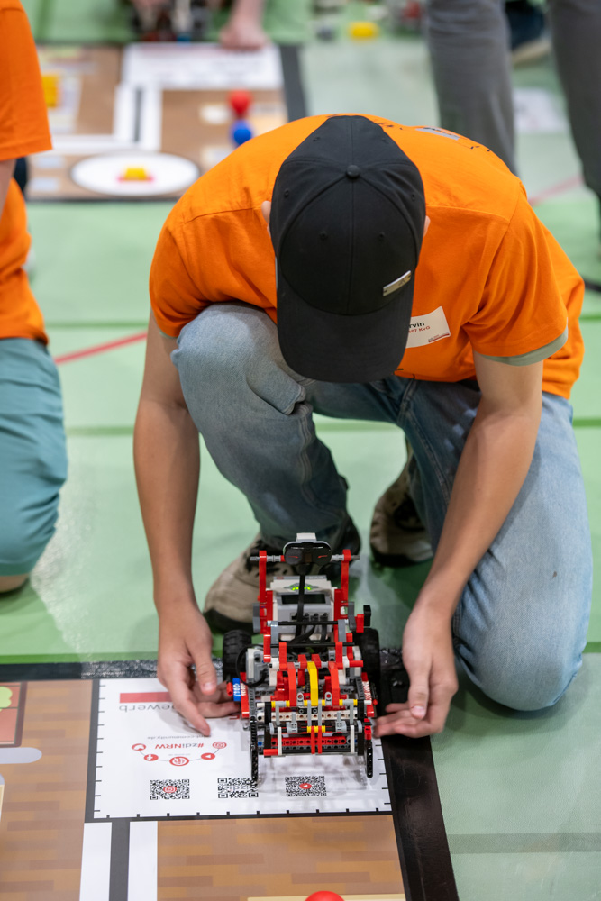 Eine Person, die ein orangefarbenes Hemd und eine schwarze Mütze trägt, justiert einen LEGO Roboter auf einer Wettbewerbsmatte bei einer Robotikveranstaltung.