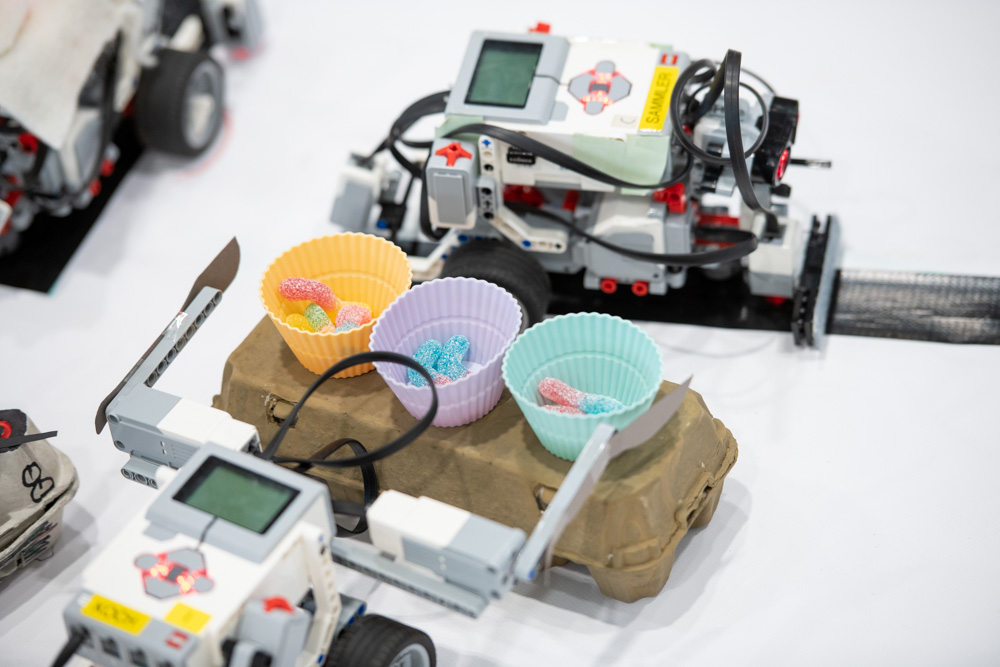 Zwei LEGO Mindstorms Roboter stehen neben drei mit Gummibonbons gefüllten Silikonbechern. Die Becher stehen auf einem Eierkarton.