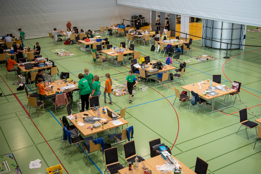 Die Menschen sind in einer großen Sporthalle versammelt, sitzen und stehen um Tische mit Laptops, Robotern und verschiedenen Geräten. Sie scheinen an einem Robotik-Wettbewerb oder -Workshop teilzunehmen.