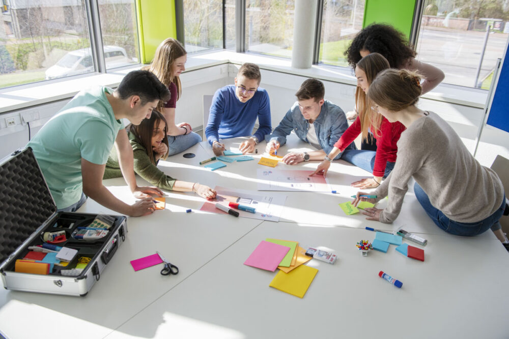 Eine Gruppe junger Erwachsener arbeitet in einem hell erleuchteten Raum um einen Tisch herum, der mit Papieren, Stiften und Büromaterialien bedeckt ist.