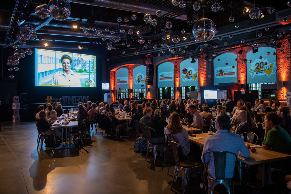Eine große Gruppe von Menschen sitzt an Tischen in einer Halle im Industriestil und sieht sich eine Präsentation auf einer großen Leinwand an.