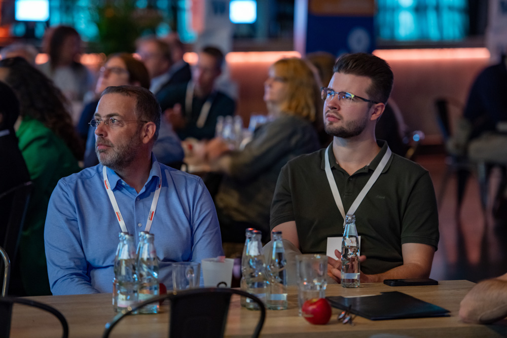 Zwei Männer mit Schlüsselbändern sitzen an einem Tisch mit Wasserflaschen, Gläsern und einem Apfel. Sie beobachten eine Veranstaltung, während andere hinter ihnen sitzen.