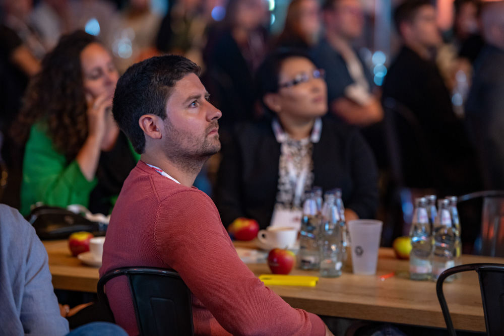 Mehrere Personen sitzen an einem Tisch und hören während einer Veranstaltung aufmerksam zu. Auf dem Tisch befinden sich Flaschen, Äpfel und Papiere.