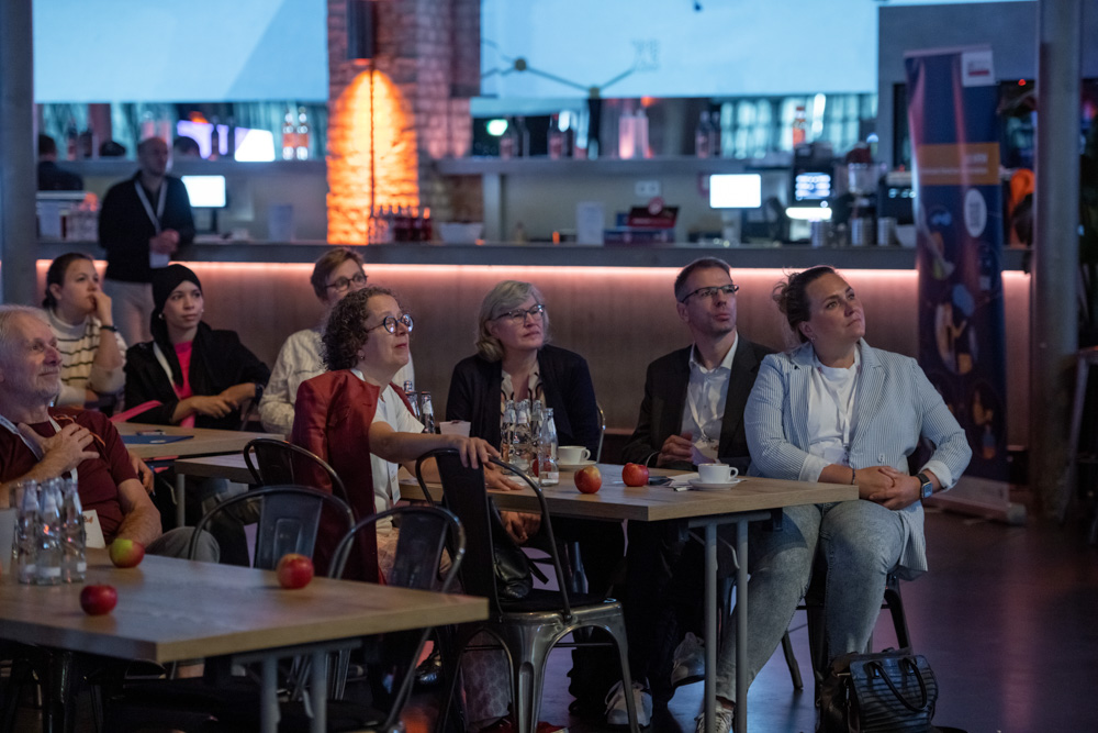Menschen sitzen an Tischen und verfolgen eine Präsentation in einem schwach beleuchteten, modernen Raum. Äpfel, Tassen und Gläser stehen auf den Tischen.