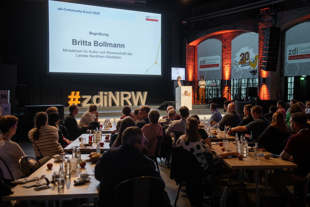 Menschen sitzen an Tischen und verfolgen eine Präsentation auf dem zdi Community Event 2023. Auf einer großen Leinwand wird Britta Bollmann begrüßt.