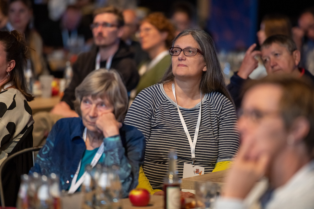 Erwachsene, die Konferenzbänder tragen, sitzen an Tischen und hören einem Redner bei einer Indoor-Veranstaltung aufmerksam zu.