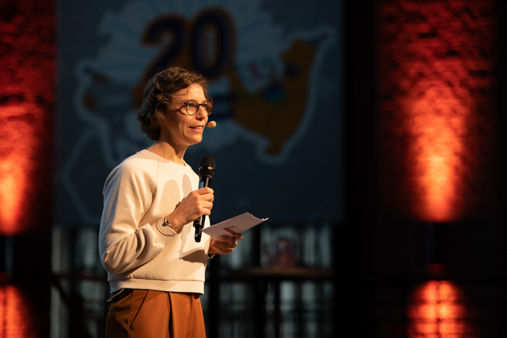 Eine Person steht auf der Bühne, hält ein Mikrofon und einen Zettel in der Hand. Im unscharfen Hintergrund sind die Zahl "20" und eine rote Beleuchtung zu sehen.