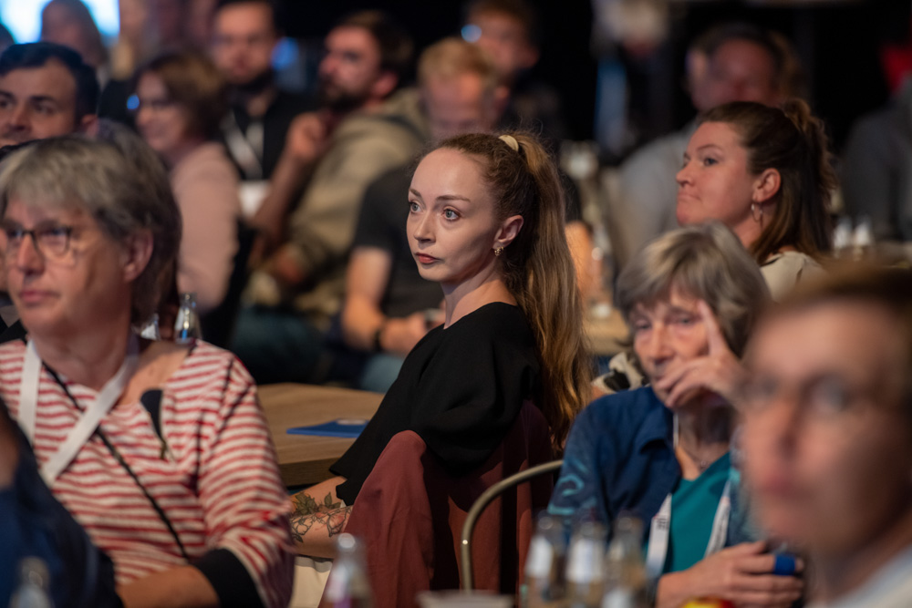 Menschen sitzen in einem Raum an Tischen und hören bei einer Veranstaltung oder Konferenz aufmerksam einem Vortrag zu.