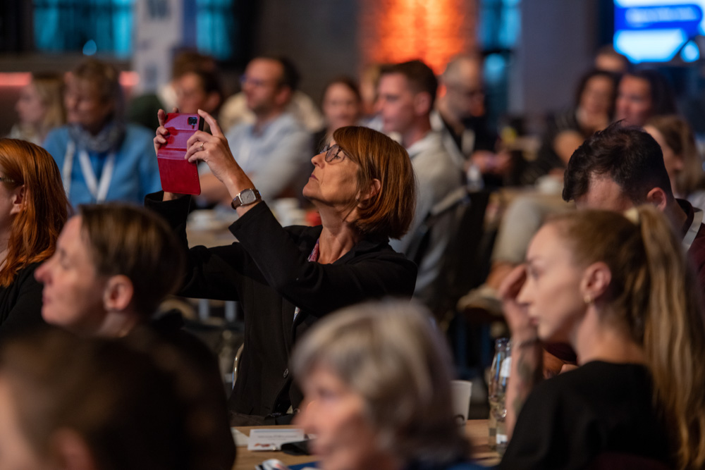 Eine Frau hebt ihr Handy, um bei einer Veranstaltung in einer Halle ein Foto zu machen, umgeben von anderen Menschen, die in der Menge sitzen.