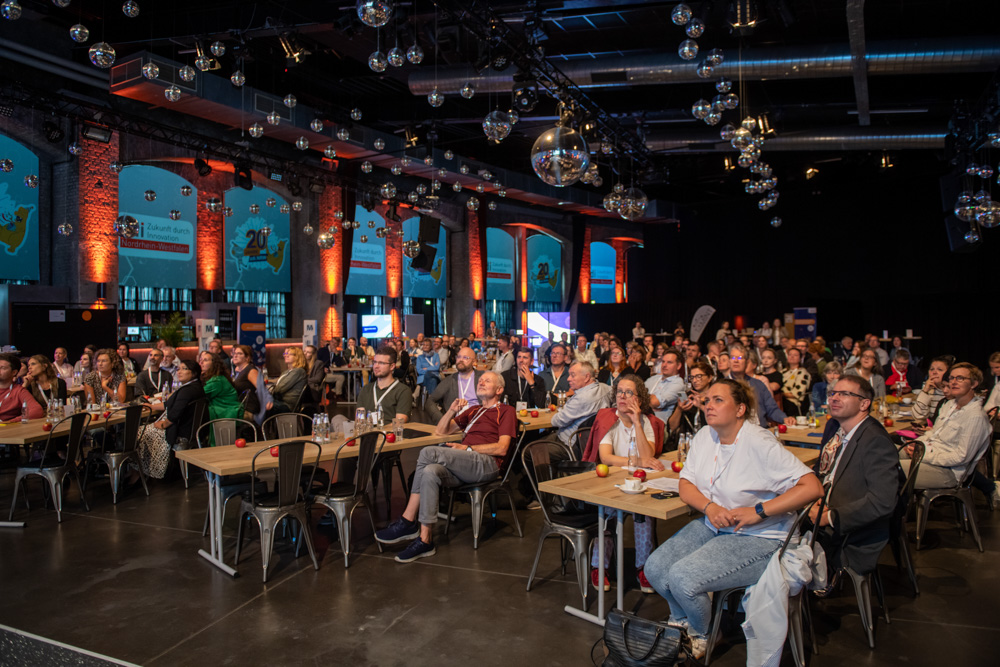 Eine große Gruppe von Menschen sitzt an Tischen in einem Veranstaltungsraum im industriellen Stil und verfolgt eine Präsentation oder Aufführung auf der Bühne.