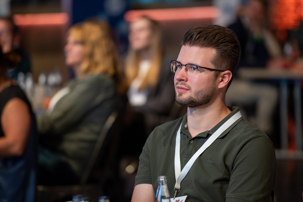 Ein Mann mit Brille und grünem Polohemd sitzt aufmerksam bei einer Indoor-Veranstaltung. Andere Personen sitzen im Hintergrund.