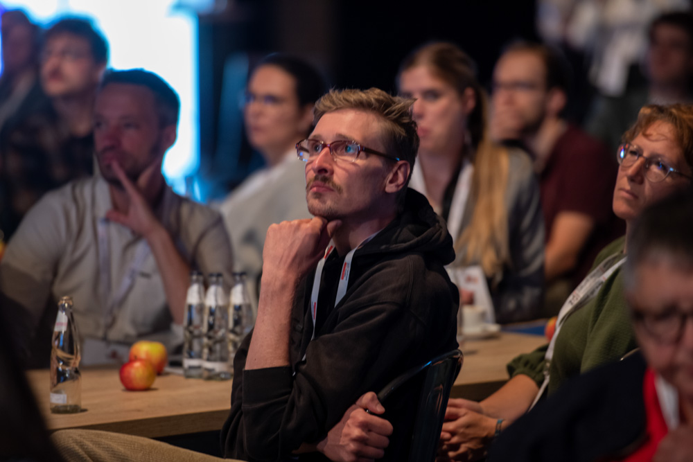 Menschen sitzen an einem Tisch und hören einem Redner bei einer Veranstaltung zu. Auf dem Tisch stehen Wasserflaschen und Äpfel.