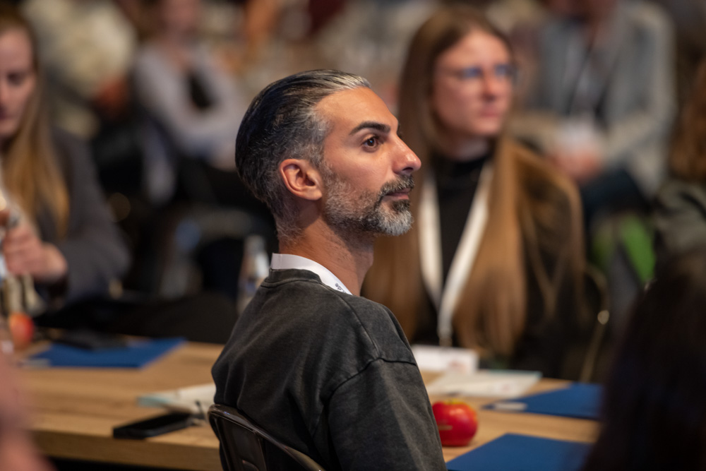 Ein Mann mit grauem Haar und Bart sitzt an einem Konferenztisch und hört aufmerksam zu. Andere Teilnehmer sitzen um ihn herum.