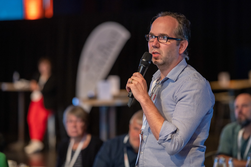 Ein Mann mit Brille spricht bei einer Indoor-Veranstaltung in ein Handmikrofon, während sitzende Personen im Hintergrund verschwommen erscheinen.