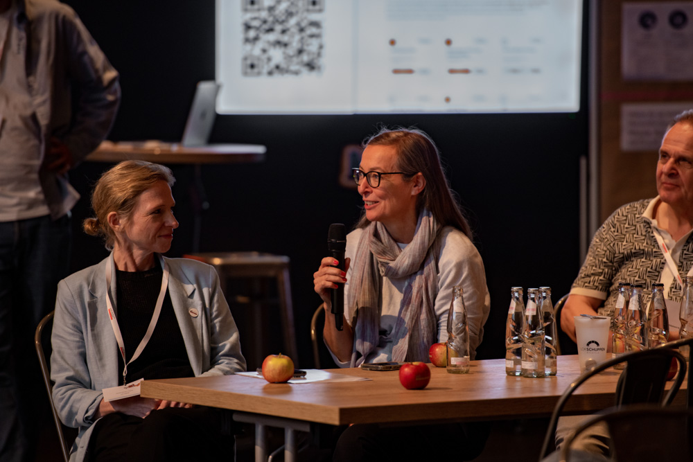 Eine Frau spricht an einem Tisch mit drei anderen Personen in ein Mikrofon. Vor ihnen liegen Äpfel und Wasserflaschen bei einer Veranstaltung in einer Halle.