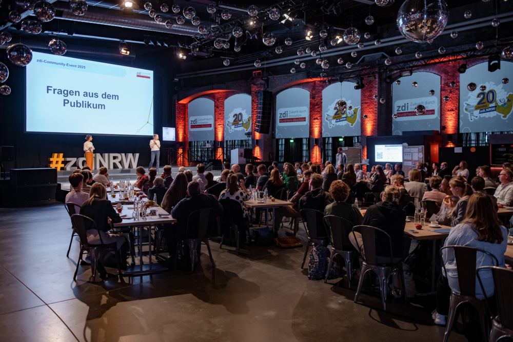 Ein großes Publikum sitzt an Tischen und blickt auf eine Bühne. In einer modernen Halle stehen zwei Redner vor einem Bildschirm mit der Aufschrift "Fragen aus dem Publikum".