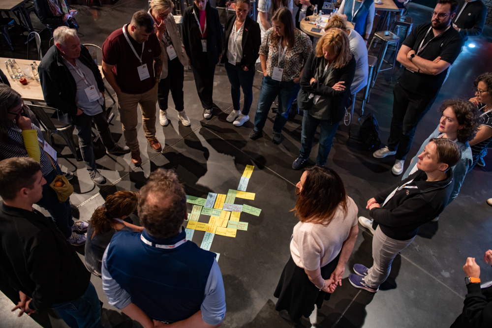 Mehrere Personen stehen in einem Kreis und betrachten bunte Klebezettel und Papiere, die in der Mitte auf dem Boden liegen.