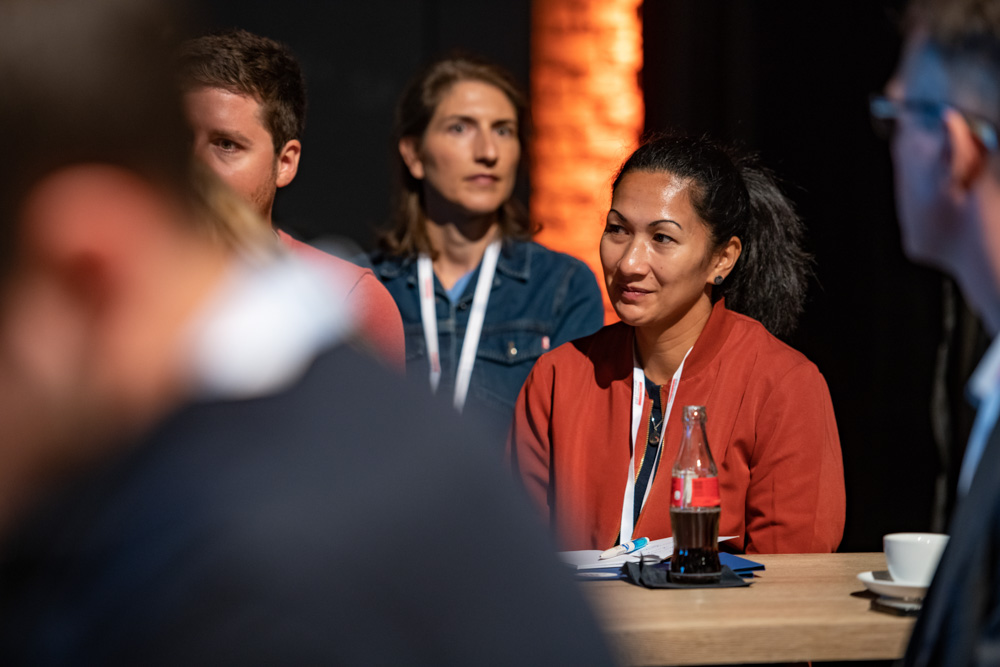 Mehrere Personen mit Konferenzbändern sitzen an einem Tisch und hören zu. Eine Frau mit roter Jacke, Notizbuch und Getränk steht im Mittelpunkt.
