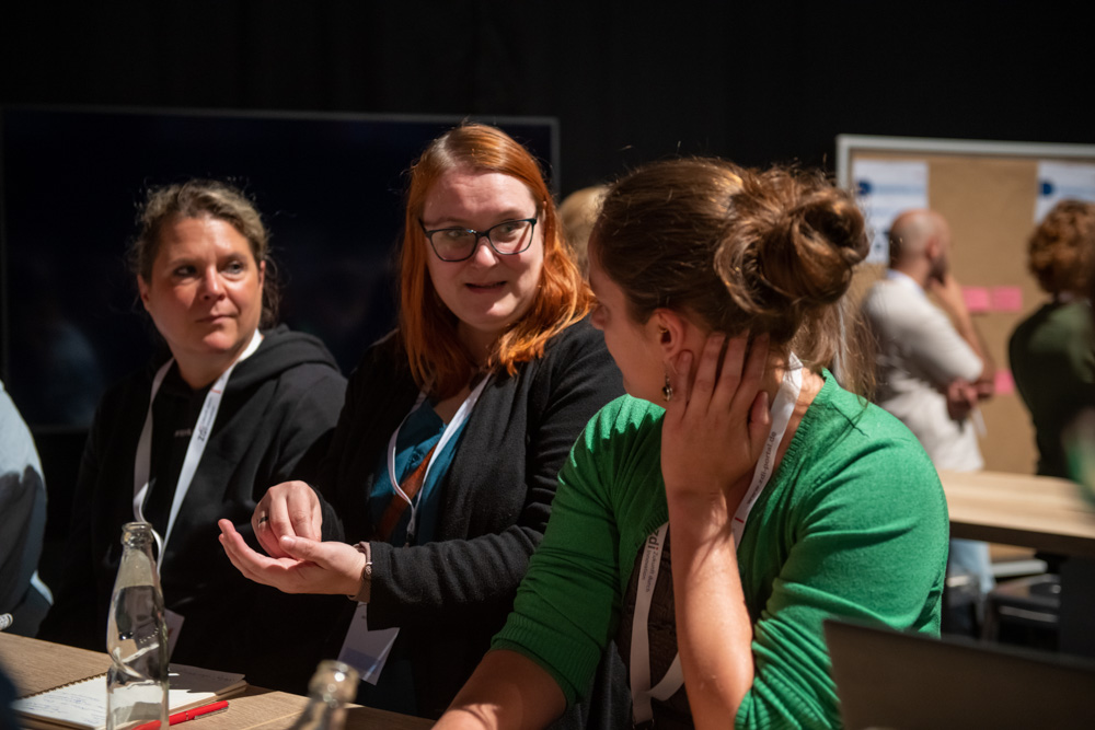 Drei Frauen sitzen an einem Tisch und unterhalten sich auf einer Konferenz oder einem Workshop. Sie tragen Schlüsselbänder und andere sind in der Nähe.