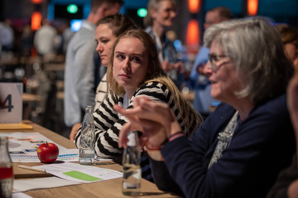 Zwei Frauen sitzen an einem Konferenztisch mit Papieren, Flaschen und einem Apfel, während sie sich unterhalten. Andere Teilnehmer sind im Hintergrund zu sehen.