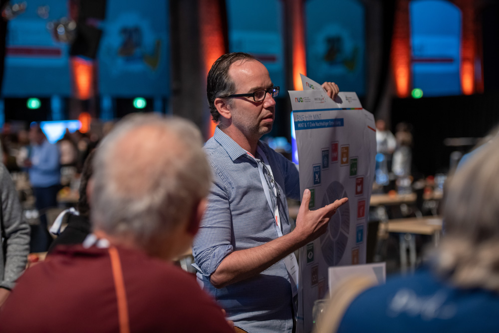 Ein Mann steht auf und präsentiert den Zuhörern einer Konferenz in einem Innenraum ein großes Diagramm.