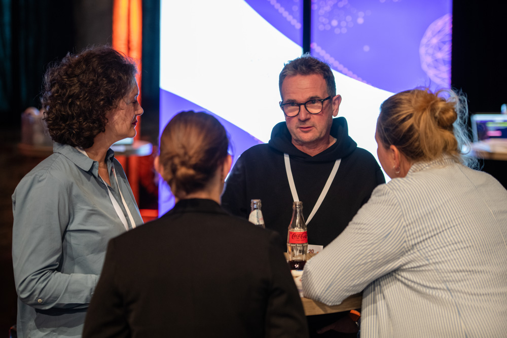 Vier Personen stehen um einen hohen Tisch herum und unterhalten sich. Auf dem Tisch stehen Getränke, und hinter ihnen befindet sich ein blau-lila Hintergrund.