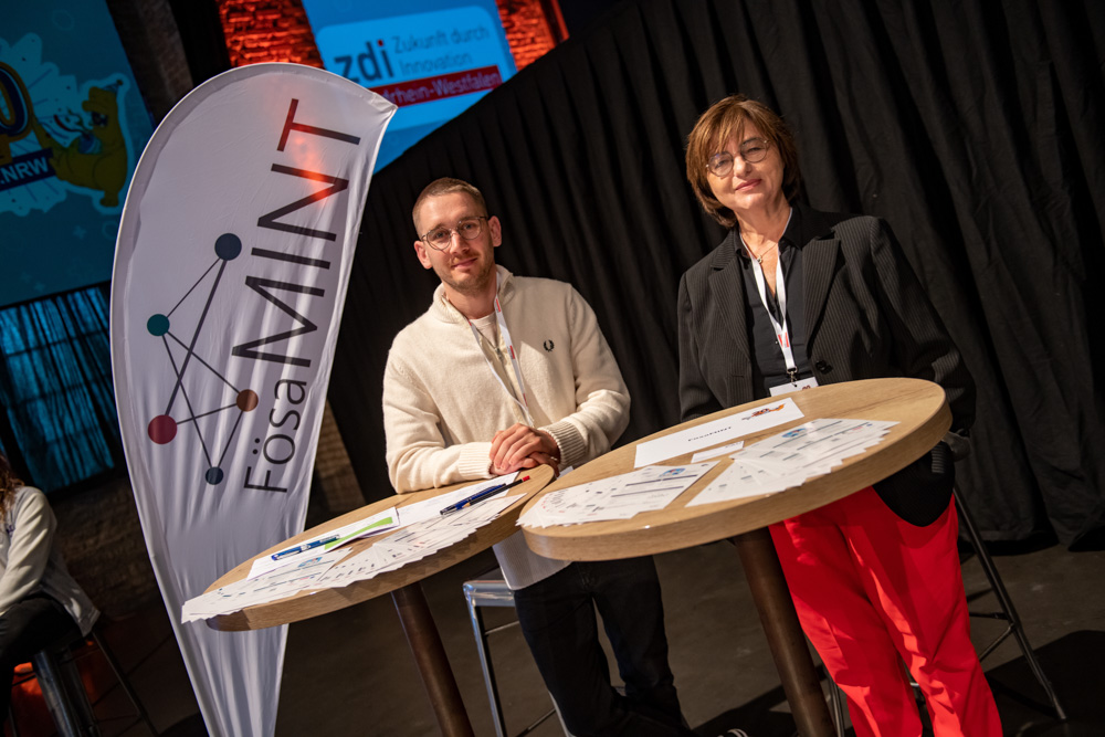 Zwei Personen stehen hinter einem hohen Tisch mit Flyern an einem FÖSA MINT-Stand. Neben ihnen steht ein FÖSA MINT-Banner im Innenbereich.