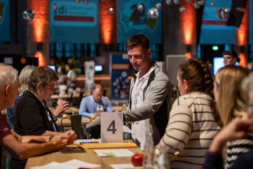 Mehrere Personen sitzen bei einer Indoor-Veranstaltung um einen mit der Zahl 4 beschrifteten Tisch und diskutieren mit Unterlagen und Laptops.