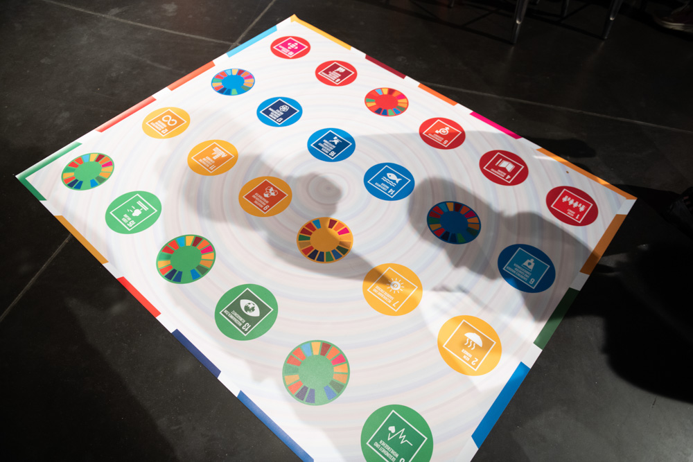 Eine bunte Tafel zeigt Symbole für die Ziele der Vereinten Nationen für nachhaltige Entwicklung, die in einem Kreis auf einem dunklen Boden angeordnet sind.