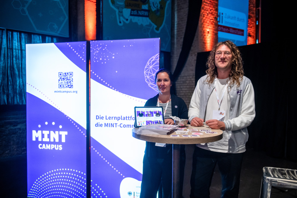 Zwei Personen stehen an einem runden Tisch mit einem Tablet und Aufklebern und werben für MINT Campus vor einem gebrandeten Indoor-Display.