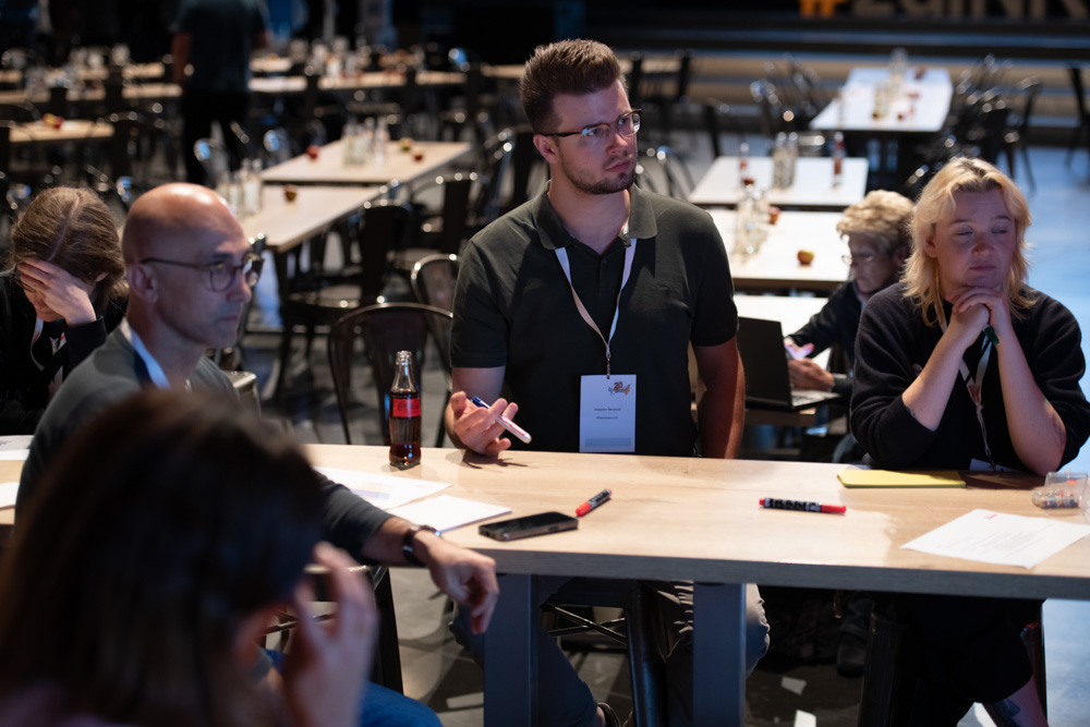 Mehrere Personen sitzen um einen Stehtisch mit Papieren und Stiften. Sie sind konzentriert und in eine Diskussion auf einer Konferenz oder einem Workshop vertieft.