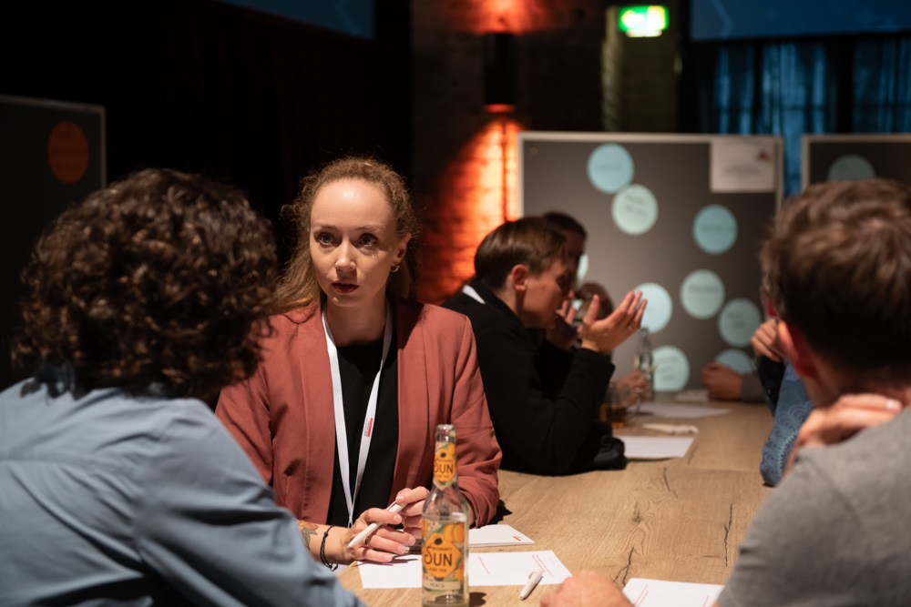 Eine Frau mit lockigem Haar und einem rosa Blazer sitzt während einer Gruppendiskussion am Tisch und hört zu. Auf dem Tisch stehen Getränke und Papiere.