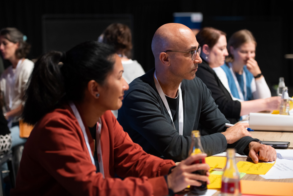 Während einer Sitzung oder eines Workshops sitzen mehrere Personen an einem Tisch und hören aufmerksam zu. Auf dem Tisch liegen Notizbücher, Stifte und Limonadenflaschen.