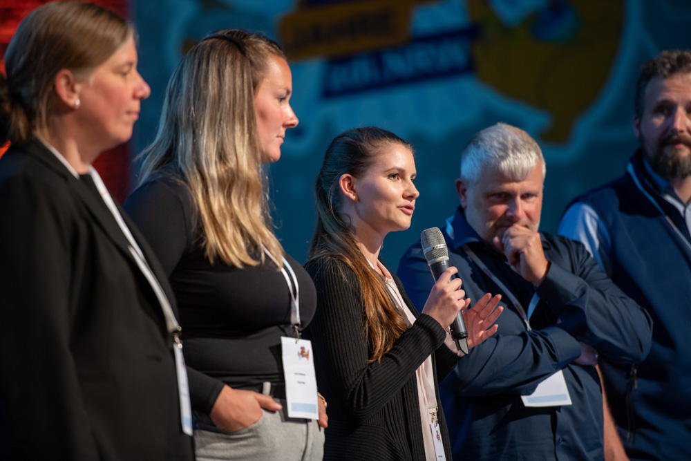 Fünf Personen stehen bei einer Veranstaltung in einer Reihe. Die Frau in der Mitte spricht in ein Mikrofon, und die anderen hören aufmerksam zu.