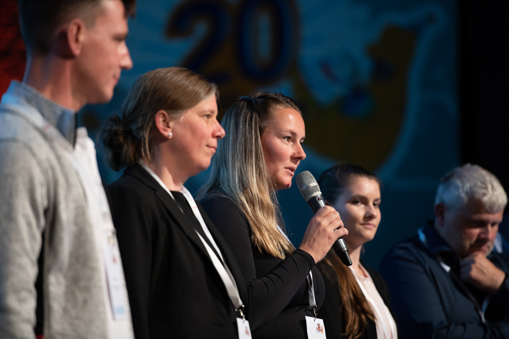 Fünf Personen stehen in einer Reihe auf der Bühne. Die dritte Person hält ein Mikrofon und spricht. Die anderen hören zu. Auf dem Hintergrund ist die Zahl 20 zu sehen.