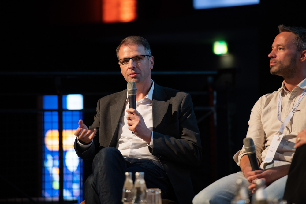 Ein Mann im Anzug spricht in ein Mikrofon, während er neben einem anderen Mann auf einem Podium sitzt. Auf dem Tisch vor ihnen stehen Wasserflaschen.