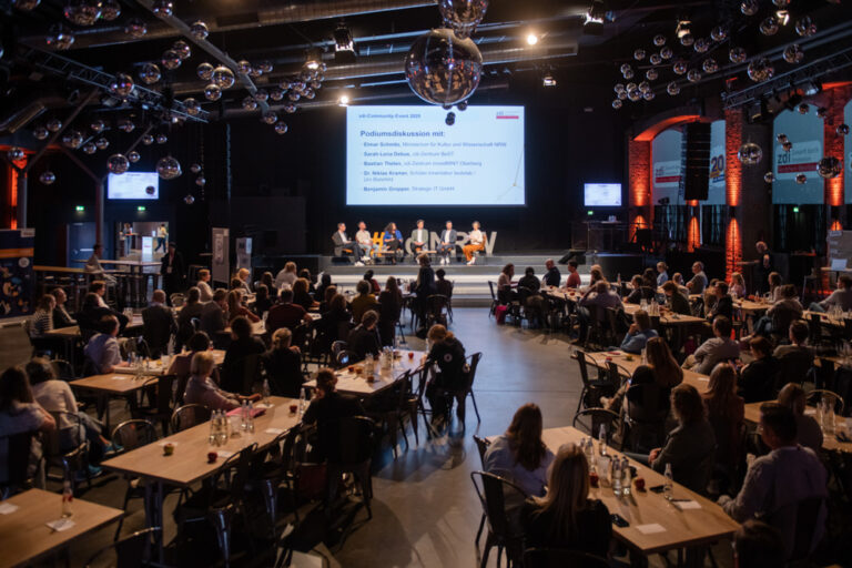 Ein großes Publikum sitzt an Tischen. Sie verfolgen eine Podiumsdiskussion auf der Bühne in einem geräumigen Veranstaltungssaal mit dekorativer Beleuchtung und einer Leinwand.
