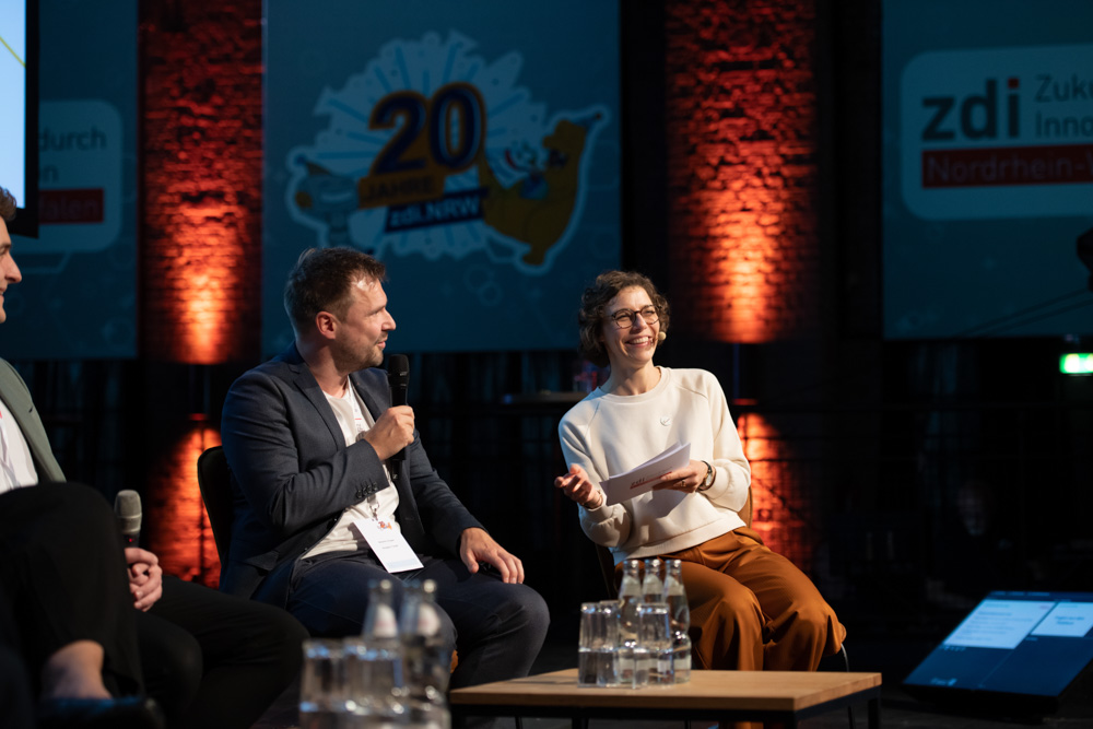 Drei Personen sitzen auf der Bühne für eine Diskussionsrunde. Eine Frau mit Papieren lächelt, während eine andere Person in ein Mikrofon spricht. Transparente und Wasserkrüge sind vorhanden.