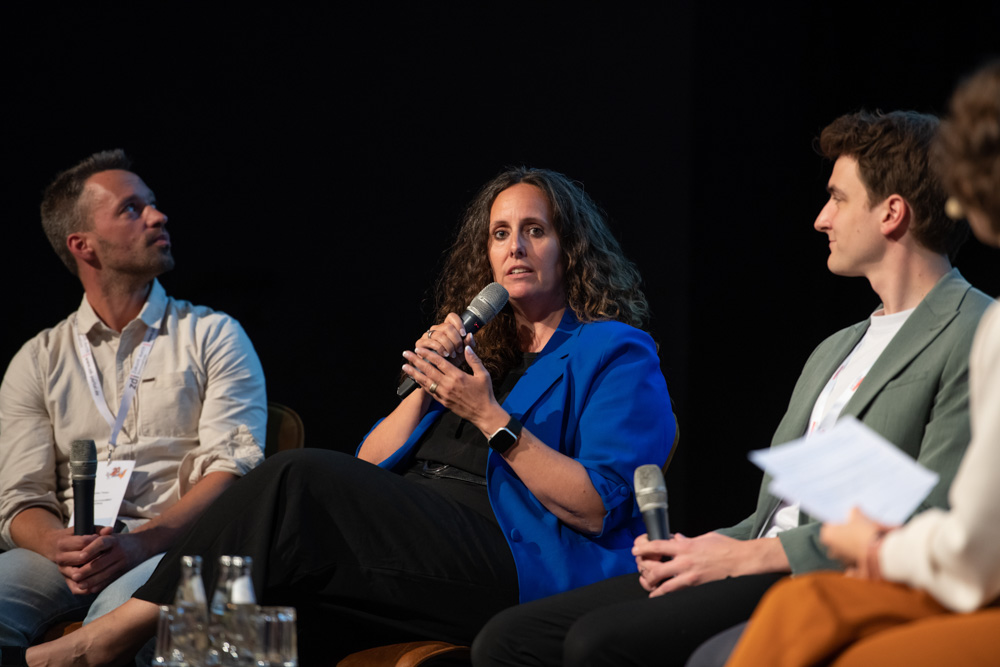 Vier Personen sitzen bei einer Podiumsdiskussion auf der Bühne. Eine Frau in einem blauen Blazer spricht in ein Mikrofon, während die anderen aufmerksam zuhören.
